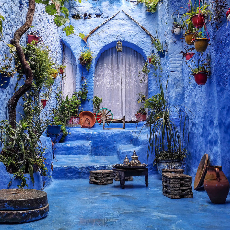 Cour intérieure typique de Chefchaouen, la célèbre ville bleue du Maroc, murs et sol peints en bleu vif, ambiance traditionnelle, pots de fleurs suspendus et au sol, plantes luxuriantes, marches menant à une porte avec rideau blanc brodé, objets artisanaux marocains comme moulin à épices et théière, décor illustrant architecture, artisanat et patrimoine marocains, idéal pour tourisme, circuits culturels, escapades photogéniques, voyages immersifs, découverte artisanat et gastronomie locale.